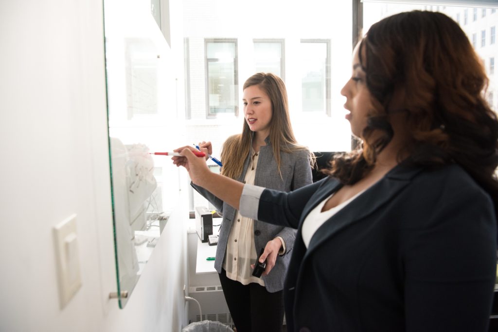 Two women writing on a glass whiteboard screen inside an office building.