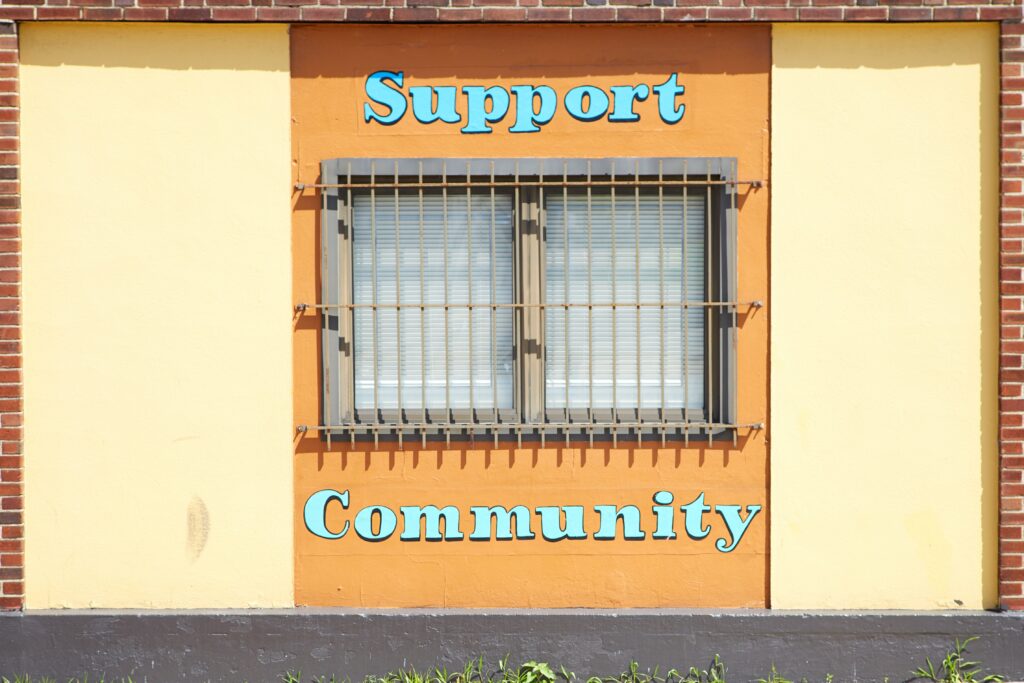 Orange painted brick wall with a barred window and blue painted lettering that says ‘Support Community