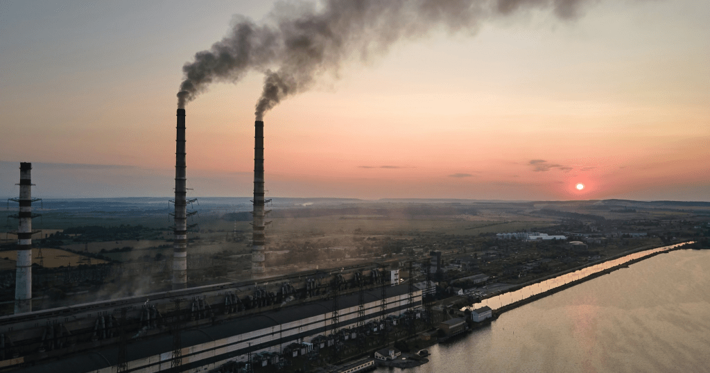 Factories producing visible emissions via a chimney stack with a sunrise in the background.