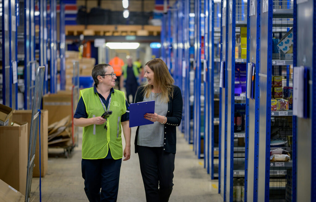 Two colleagues walk through a warehouse, both smiling. One is in a high-vis vest, the other is holding a clipboard.