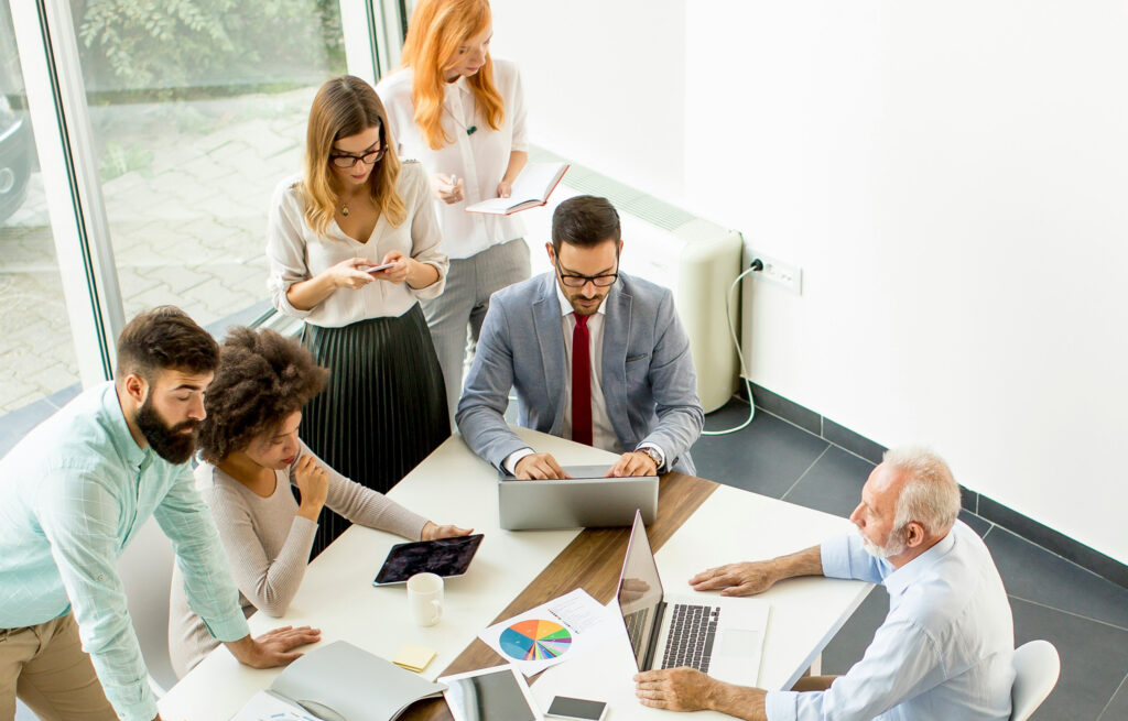 Group of people of varying ages and ethnicities sit around a business table having a meeting.