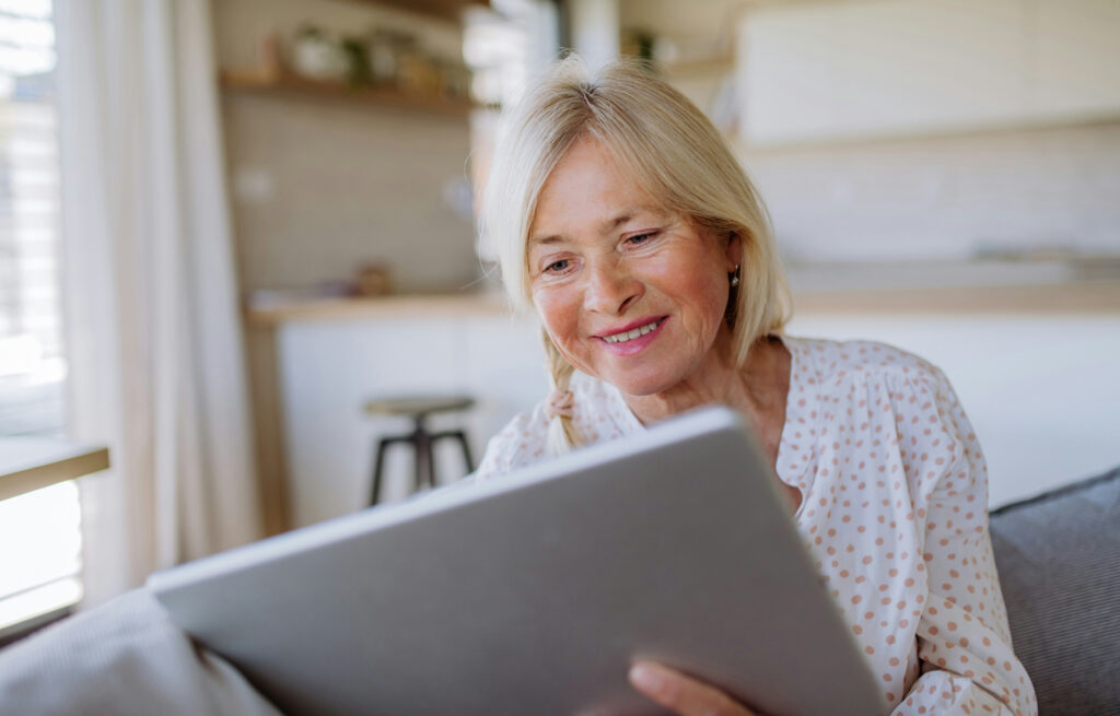 Older female sitting on a sofa, looking at information on a tablet screen.