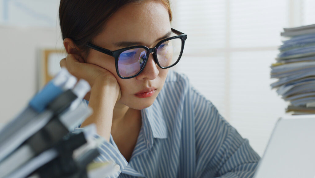 Woman in glasses surrounded by paper and concentrating hard while looking at her computer screen.