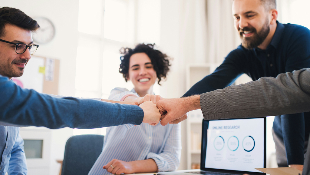 Woman smiling. She puts her fist in a fist high-five huddle with other peoples hands suggesting they've just set meeting goals and objectives.